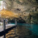 Das Ostauge der Cenote Dos Ojos: ein Kalksteingewölbe voller Stalaktiten über türkisfarbenem Wasser, links ein Holzsteg und hinten Schwimmer
