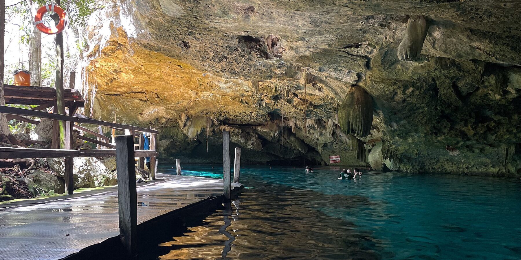 Das Ostauge der Cenote Dos Ojos: ein Kalksteingewölbe voller Stalaktiten über türkisfarbenem Wasser, links ein Holzsteg und hinten Schwimmer