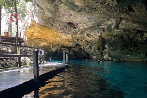 Das Ostauge der Cenote Dos Ojos: ein Kalksteingewölbe voller Stalaktiten über türkisfarbenem Wasser, links ein Holzsteg und hinten Schwimmer