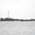 Ein schlanker weißer Leuchtturm mit roter Tür steht auf der flachen, felsigen Insel Formigão, dunkle Felsen mit braunem Algensaum, über einem grauen, aufgewühlten Meer unter bedecktem Himmel