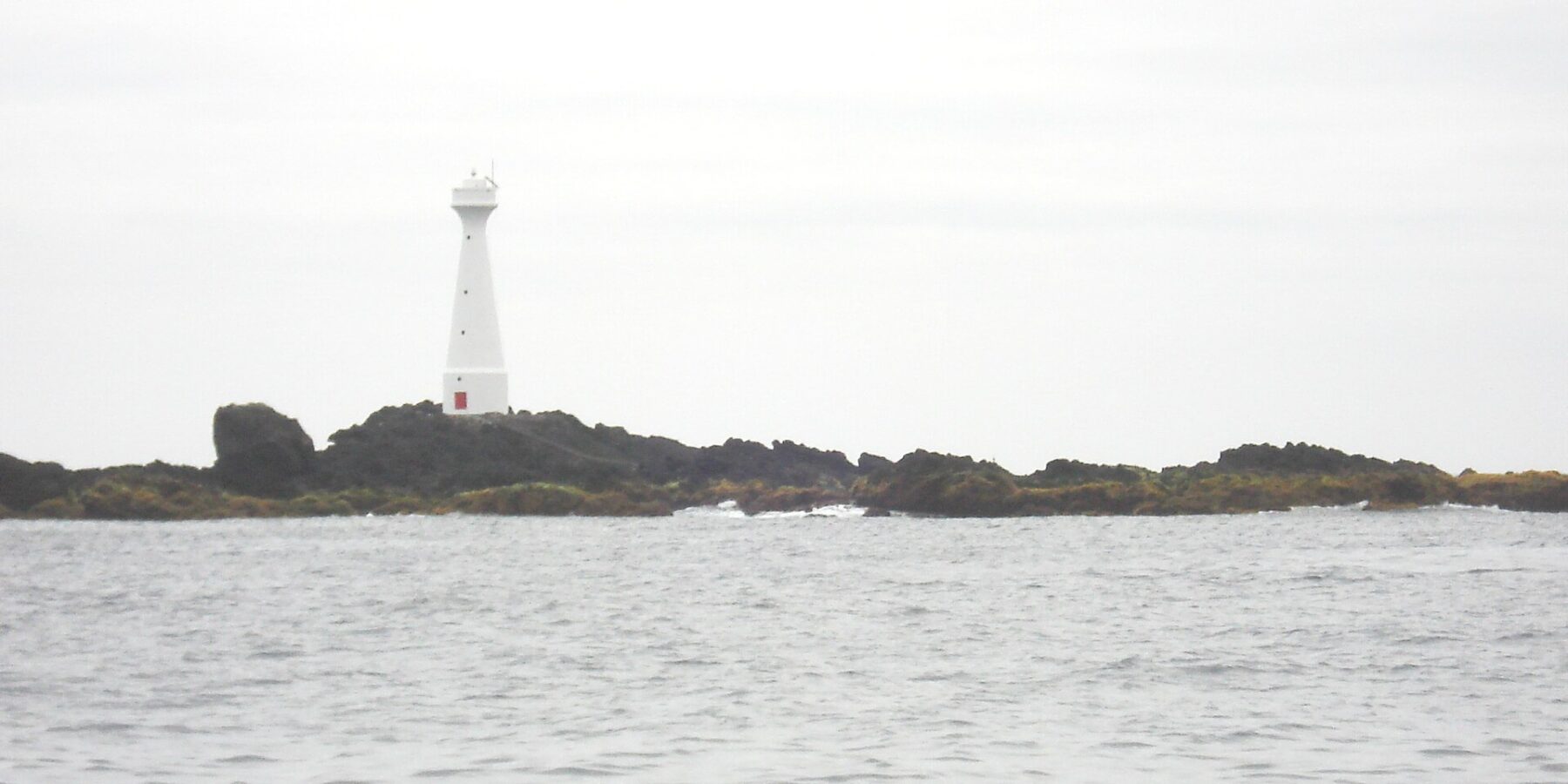 Ein schlanker weißer Leuchtturm mit roter Tür steht auf der flachen, felsigen Insel Formigão, dunkle Felsen mit braunem Algensaum, über einem grauen, aufgewühlten Meer unter bedecktem Himmel