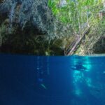 Halb über, halb unter Wasser am Eingang der Cenote The Pit: oben das Kalksteingewölbe, die Holztreppe und der Dschungel, unten zwei Taucher im tiefblauen Wasser