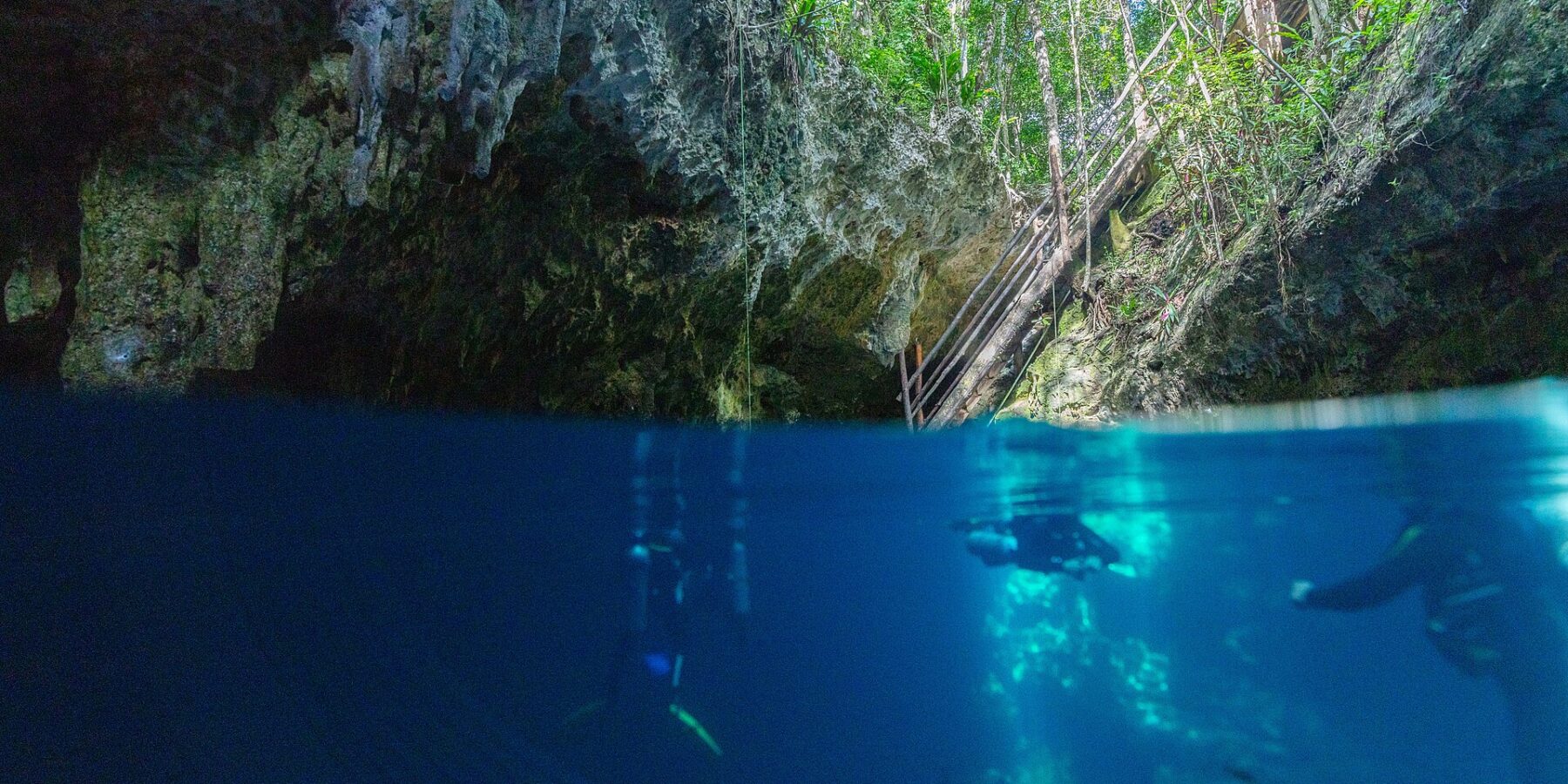 Halb über, halb unter Wasser am Eingang der Cenote The Pit: oben das Kalksteingewölbe, die Holztreppe und der Dschungel, unten zwei Taucher im tiefblauen Wasser