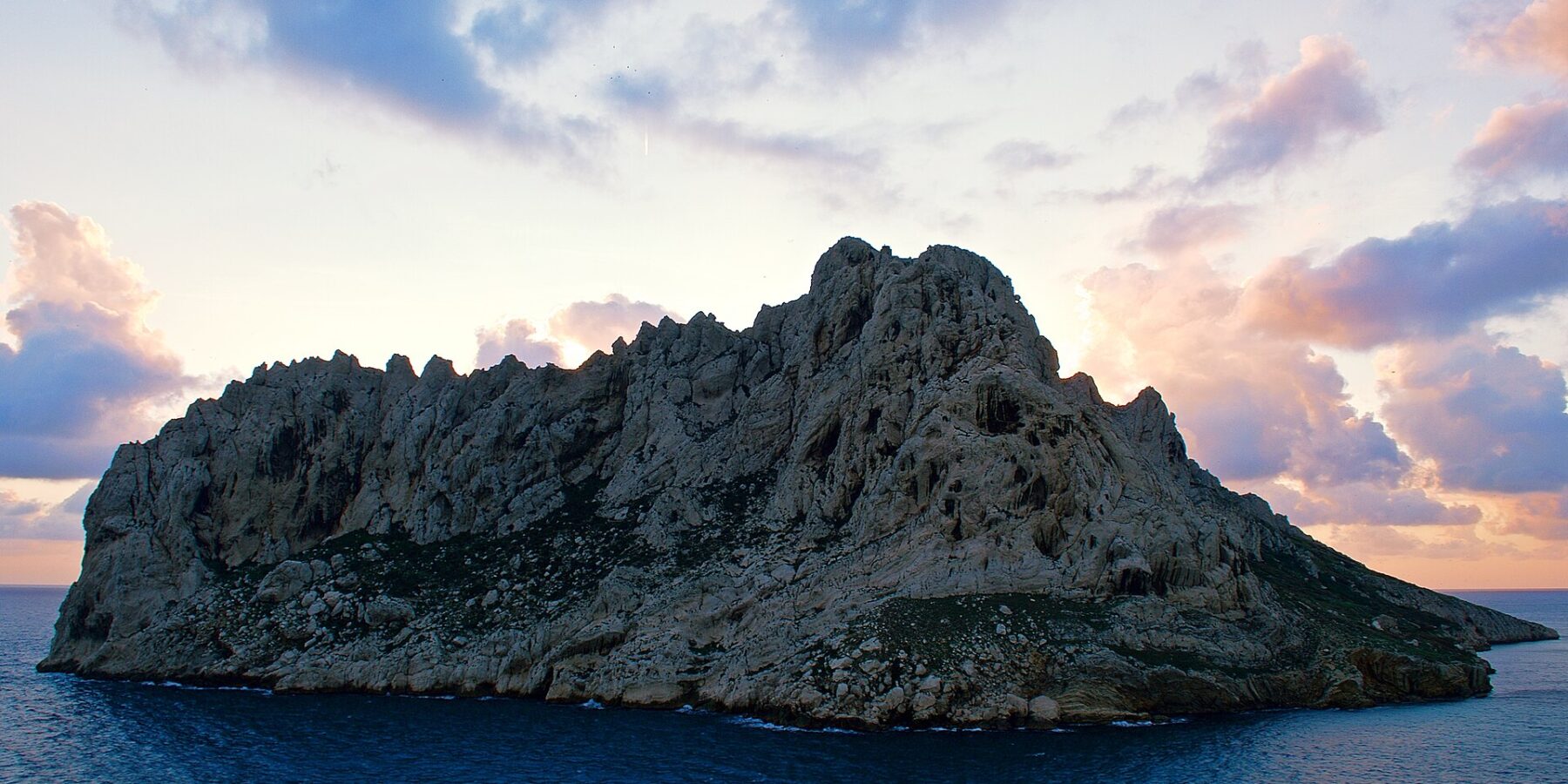 Die Île Maïre, ein heller Kalksteinblock mit zerklüfteten Graten, hebt sich im Gegenlicht vor einem Sonnenuntergangshimmel mit rosa und violetten Wolken ab, über einem dunkelblauen Meer.