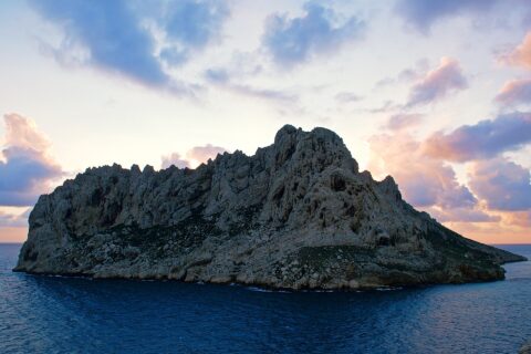 Die Île Maïre, ein heller Kalksteinblock mit zerklüfteten Graten, hebt sich im Gegenlicht vor einem Sonnenuntergangshimmel mit rosa und violetten Wolken ab, über einem dunkelblauen Meer.