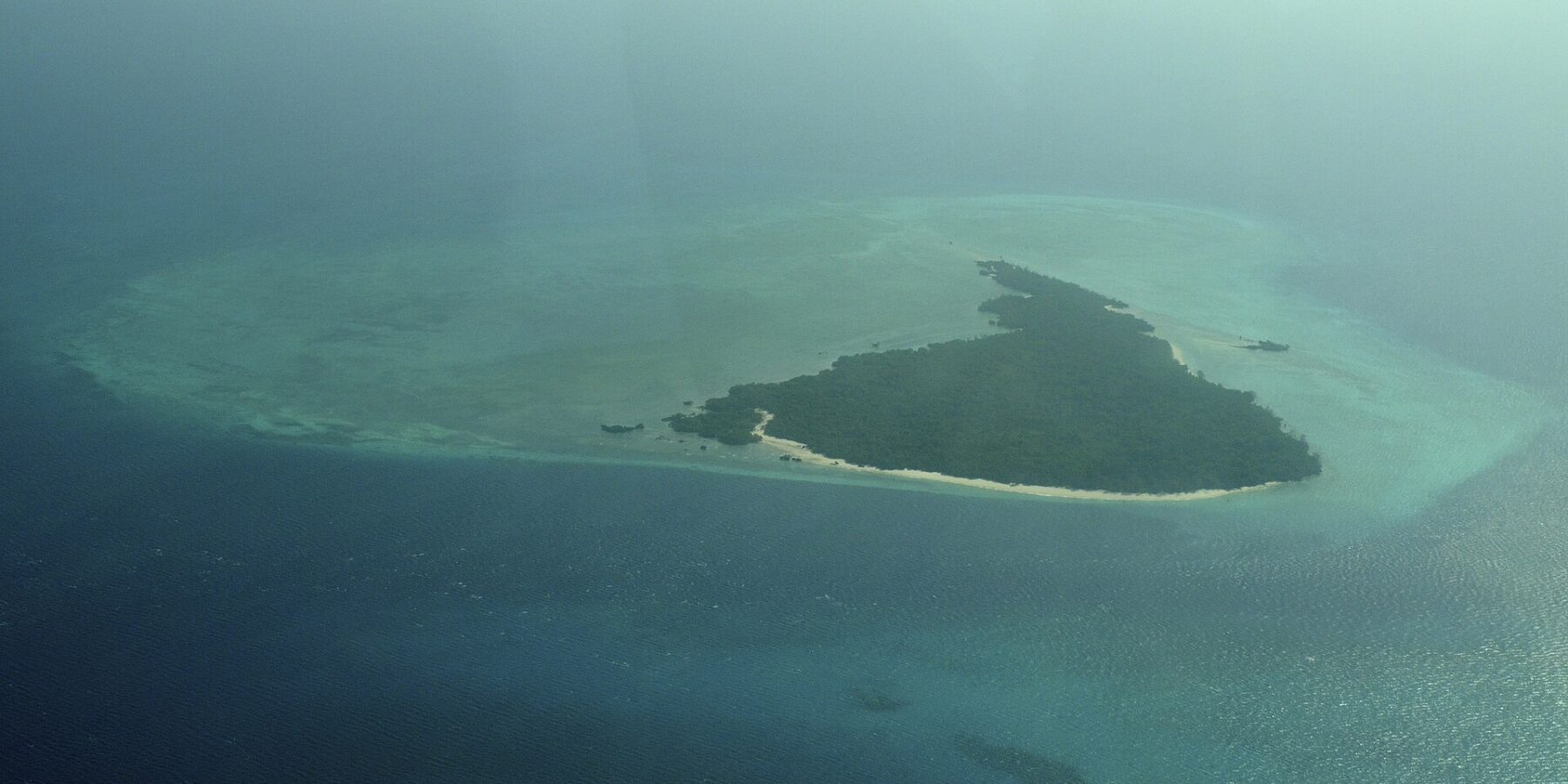 Die Insel Misali aus dem Flugzeug, ein Dreieck aus Wald mit weißem Strand, auf einem türkisen Riffdach inmitten dunkelblauen Meeres, im Dunst