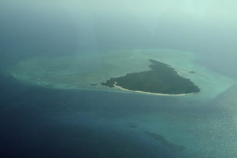 Die Insel Misali aus dem Flugzeug, ein Dreieck aus Wald mit weißem Strand, auf einem türkisen Riffdach inmitten dunkelblauen Meeres, im Dunst
