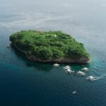 Luftbild der Insel Pescador, ein grün bewachsener Felsen mit weißem Leuchtturm, umgeben von rund zehn Tauchbancas auf dunkelblauem Meer