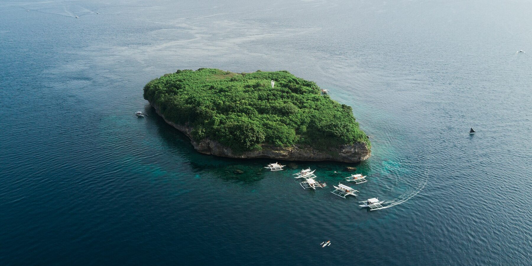 Luftbild der Insel Pescador, ein grün bewachsener Felsen mit weißem Leuchtturm, umgeben von rund zehn Tauchbancas auf dunkelblauem Meer