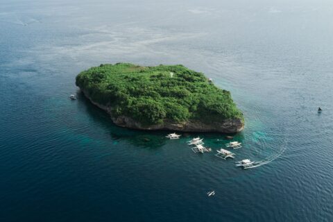 Luftbild der Insel Pescador, ein grün bewachsener Felsen mit weißem Leuchtturm, umgeben von rund zehn Tauchbancas auf dunkelblauem Meer