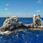 Die Roca Partida aus der Drohnenperspektive: ein von Vögeln weiß gefärbter Felsen, in zwei Spitzen gespalten, umspült von dunkelblauem Meer unter blauem Himmel mit Wolken