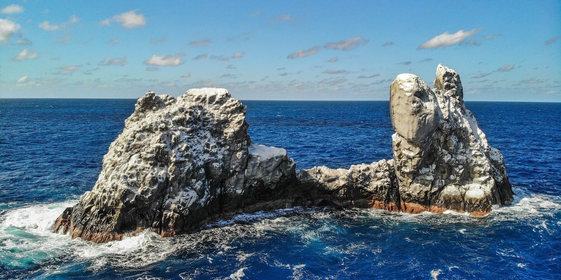 Die Roca Partida aus der Drohnenperspektive: ein von Vögeln weiß gefärbter Felsen, in zwei Spitzen gespalten, umspült von dunkelblauem Meer unter blauem Himmel mit Wolken