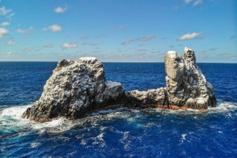 Die Roca Partida aus der Drohnenperspektive: ein von Vögeln weiß gefärbter Felsen, in zwei Spitzen gespalten, umspült von dunkelblauem Meer unter blauem Himmel mit Wolken