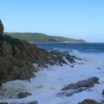 Wellen brechen sich in weißer Gischt an den Granitfelsen von L'Escalet im Vordergrund, vor einem aufgewühlten, dunkelblauen Meer, in der Ferne die bewaldete Küste des Cap Camarat mit seinem weißen Leuchtturm.