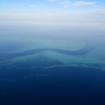 Luftbild eines dunkelblauen, S-förmigen Kanals, der sich zwischen den türkisfarbenen Riffdächern des Barriereriffs hindurchschlängelt, einige kleine weiße Boote im Pass