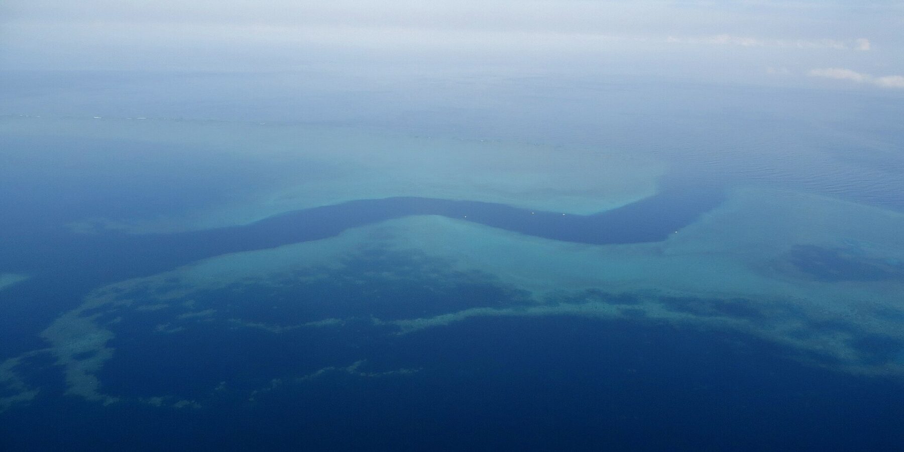 Luftbild eines dunkelblauen, S-förmigen Kanals, der sich zwischen den türkisfarbenen Riffdächern des Barriereriffs hindurchschlängelt, einige kleine weiße Boote im Pass
