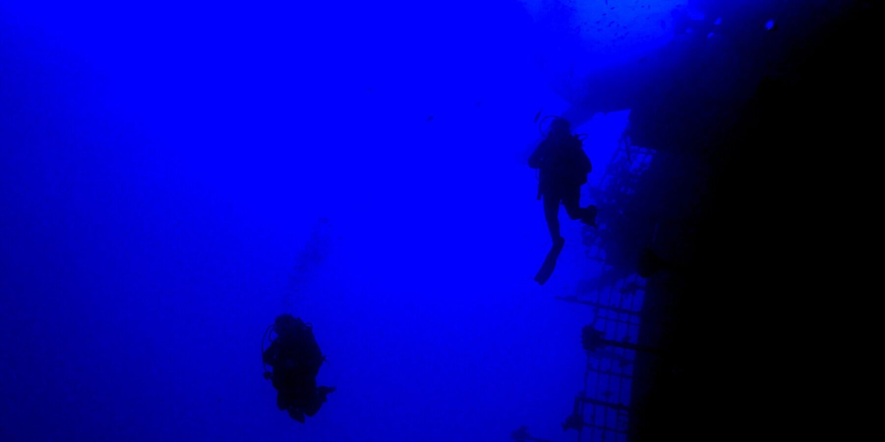 Im tiefen Blau gleiten zwei Taucher als Silhouetten an der Reling am Heck der Salem Express entlang, rechts das geneigte Deck