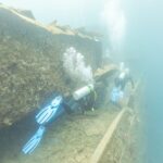 Zwei Taucher schwimmen auf dem Deck der Thistlegorm einen Gang entlang, in milchig blauem Wasser