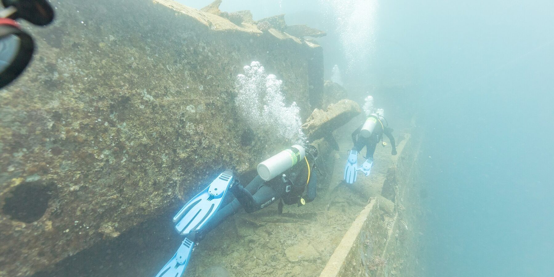 Zwei Taucher schwimmen auf dem Deck der Thistlegorm einen Gang entlang, in milchig blauem Wasser