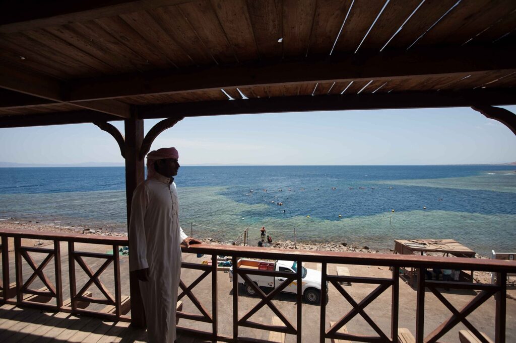 Von einer Holzterrasse aus blickt ein Mann in Galabija auf das Becken des Blue Hole, gesäumt von hellem Riff, in dem Badende schwimmen