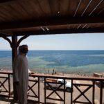 Von einer Holzterrasse aus blickt ein Mann in Galabija auf das Becken des Blue Hole, gesäumt von hellem Riff, in dem Badende schwimmen