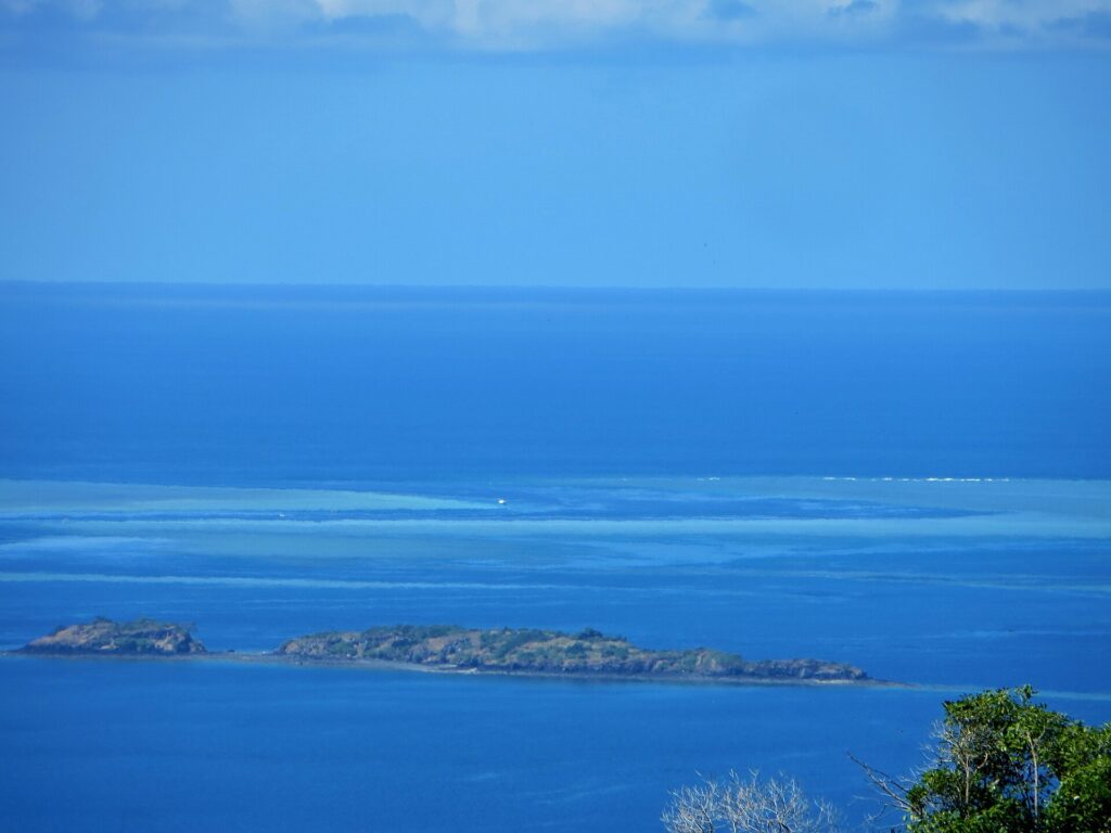 Blick von oben auf die blaue Lagune, zwei felsige Inselchen im Vordergrund und in der Ferne die hellen Riffdächer des Barriereriffs, wo ein weißes Boot im Pass liegt