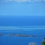 Blick von oben auf die blaue Lagune, zwei felsige Inselchen im Vordergrund und in der Ferne die hellen Riffdächer des Barriereriffs, wo ein weißes Boot im Pass liegt