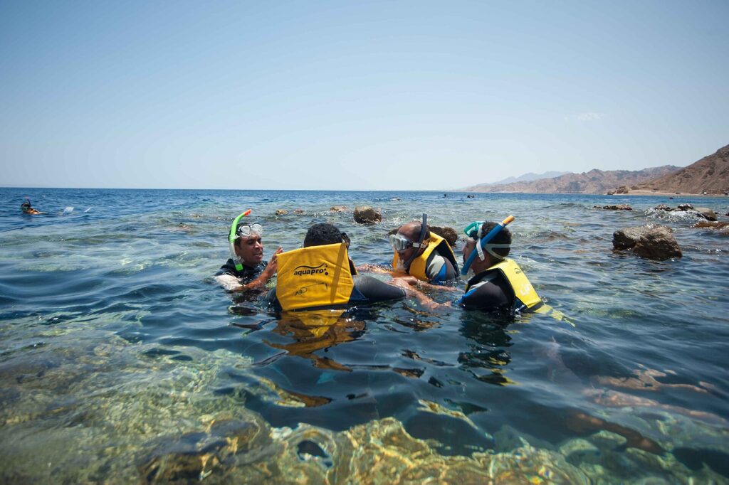 Schwimmer mit gelben Westen, Masken und Schnorcheln im klaren Wasser des Blue Hole, dahinter die Berge des Sinai